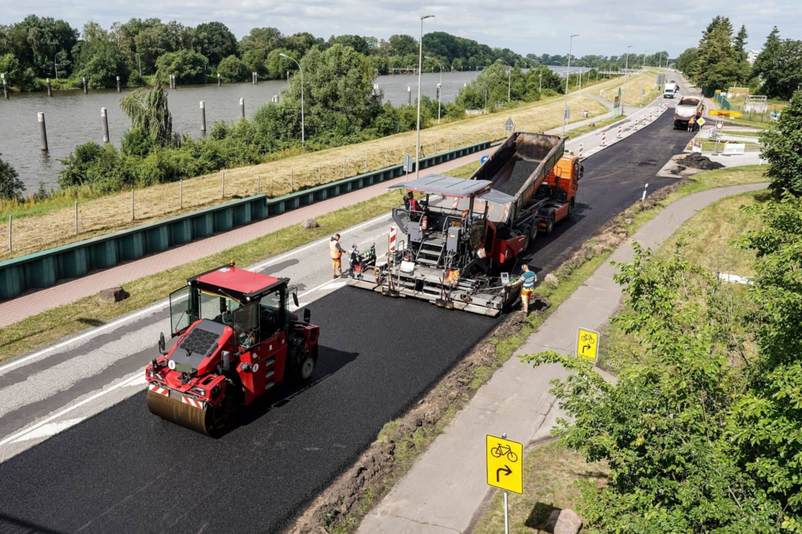 An asphalt paver laying a fresh lane, fed by a dump truck, with a roller compacting the new mat behind it.