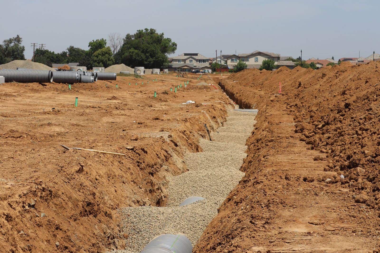 A drainage trench with pipe bedded in crushed stone on a new residential site, with more pipe and manhole sections staged nearby.